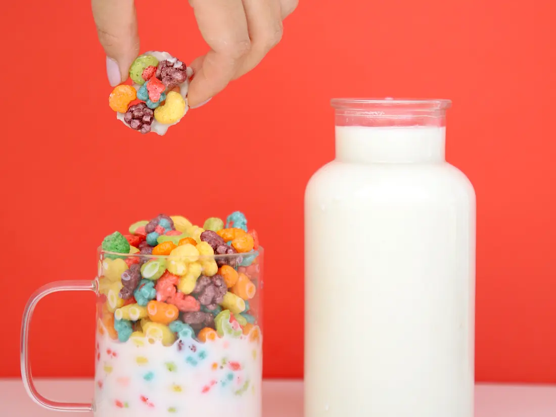 Una mano sostiene un bite de cereal sobre un vaso de cereal y una botella de leche